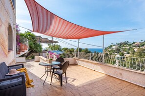 En balkon med et bord og stole, et rødt skyggesejl, udsigt til havet og en bygning med balkoner.