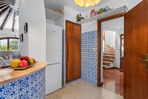 A kitchen with a fruit basket, white appliances, blue patterned tiles, and a wooden door. - Villa Axel - PlusHolidays (Calpe)