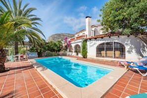 A swimming pool with a tiled deck, a white building with arches, and a mountain in the background.