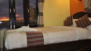 Quarto de hotel com duas camas, uma mesa de cabeceira, uma luminária e vista para o pôr do sol pela janela.