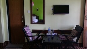 Quarto com porta de madeira, televisão fixada na parede, duas cadeiras com almofadas estampadas, uma mesa pequena com uma garrafa de água e uma chaleira, e um espelho refletindo parte do quarto.