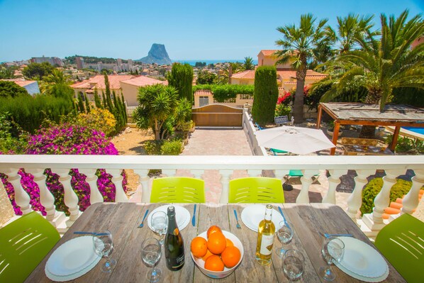 Un balcón con una mesa dispuesta para comer, con vista a un paisaje costero que incluye edificios y una montaña.
