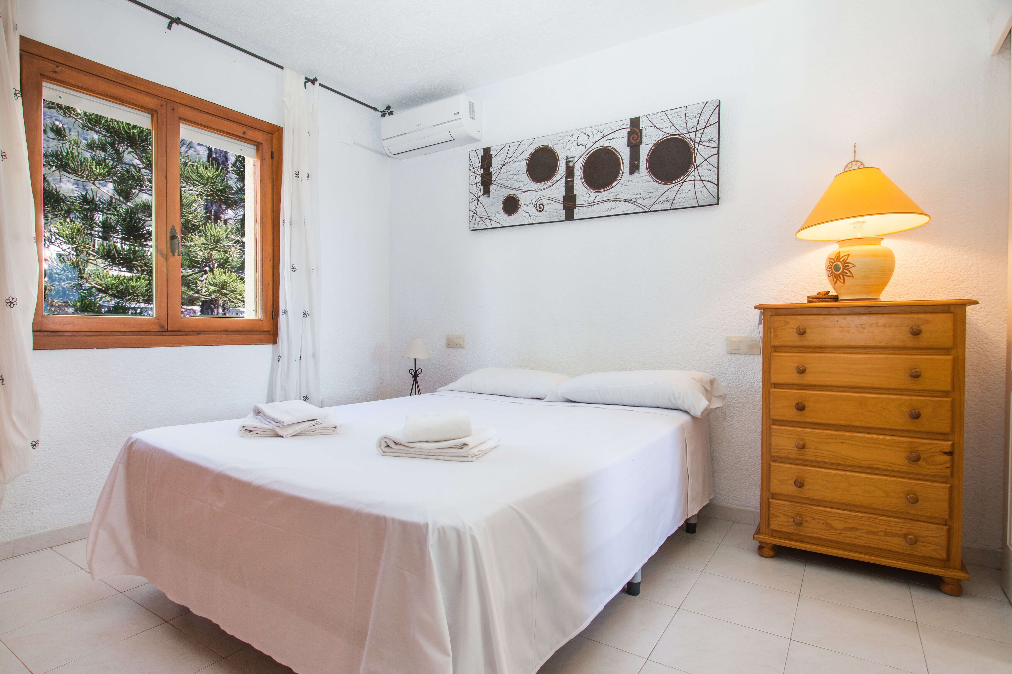 A bedroom with a bed, a dresser, a window with curtains, and a wall-mounted air conditioner.