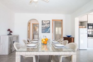 A modern kitchen with white cabinets, a dining area with a glass table and wicker chairs, and a painting of a boat on the wall. - Villa Erik - PlusHolidays (Calpe)