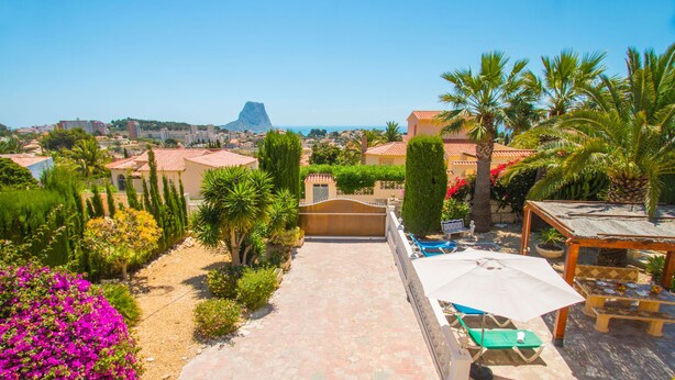Ein Gartenweg führt zu einer Terrasse mit Gartenmöbeln und Blick auf eine Küstenstadt.
