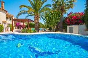 En swimmingpool med krystalklart, blå vand, omgivet af palmer og en pergola.
