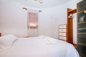A neatly made bed with a white duvet, a wooden wardrobe with a mirror, and a small bedside table with a lamp.