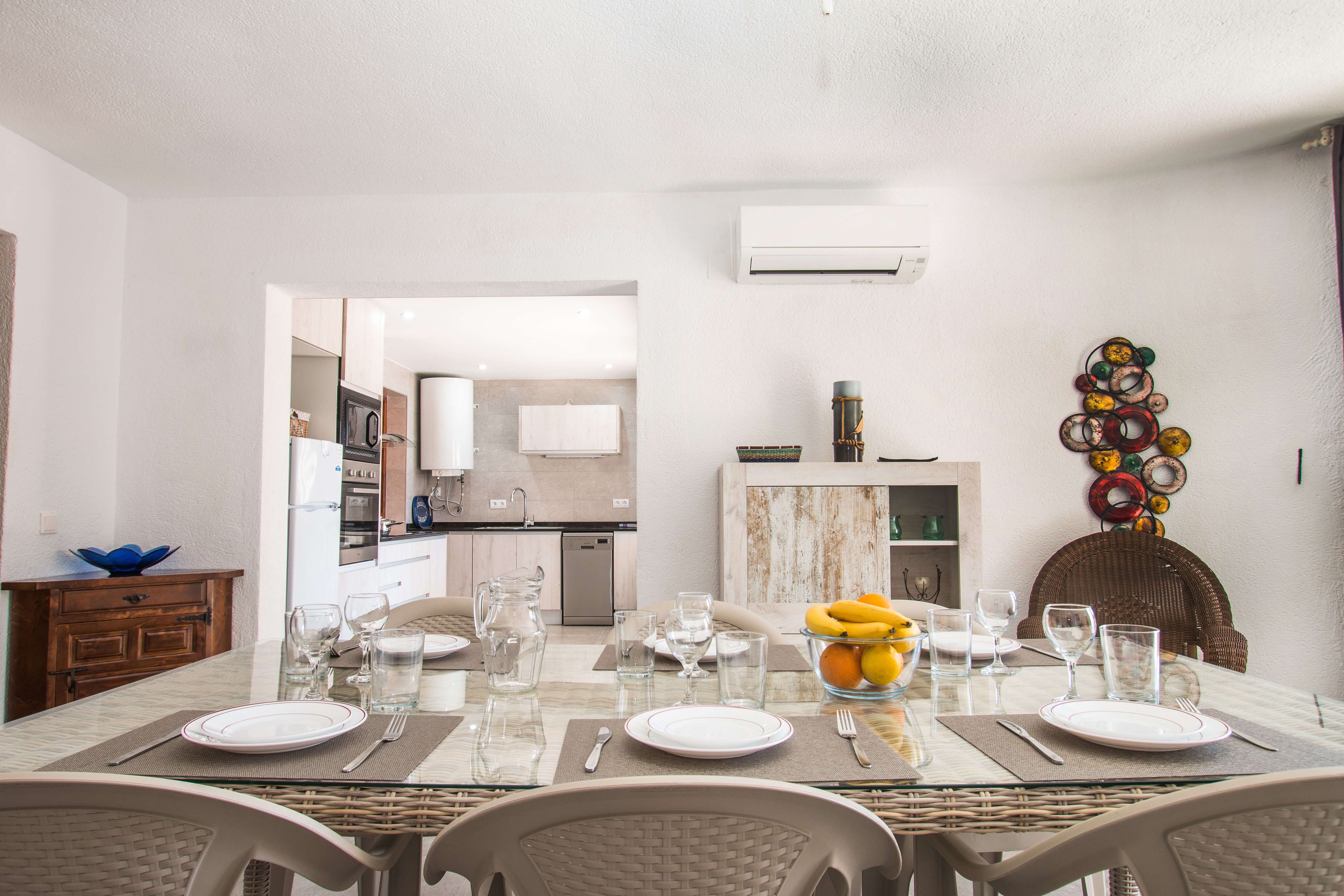 A dining area with a table set for four, a kitchen in the background, and a decorative wall piece.