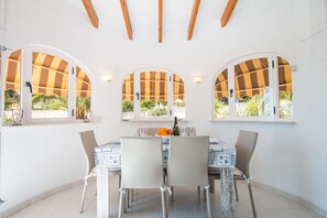 Un comedor con una mesa dispuesta para cuatro personas, rodeado de ventanales de arco, toldos a rayas, paredes blancas y un suelo de baldosas.