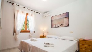 Ein Schlafzimmer mit einem Bett, zwei Nachttischen, einer Wand-Klimaanlage und einem Gemälde einer Küstenlandschaft.