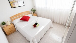 A neatly made bed with a red pillow, a small bedside table with a lamp, a potted plant, and a framed picture on the wall. - AirCon Apartment in front of the beach (La Pineda)