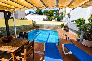 A swimming pool with a tiled deck, a wooden table, and two lounge chairs. - VILLA BUDA  (Salou)