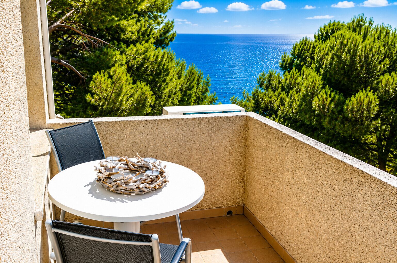 Een balkon met een ronde tafel en stoelen, met uitzicht op de kust, bomen en de zee.