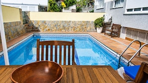 A pool with a stone wall, a wooden chair, and a wooden table with a bowl. - VILLA BUDA  (Salou)