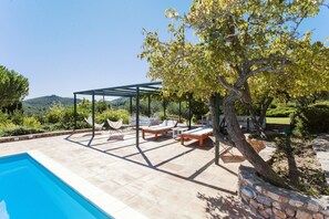 A poolside area with a pergola, lounge chairs, and a view of the hills.
