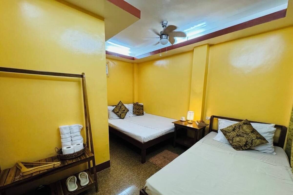 A room with two beds, a ceiling fan, a small table with a lamp, and a shelf with towels and slippers.