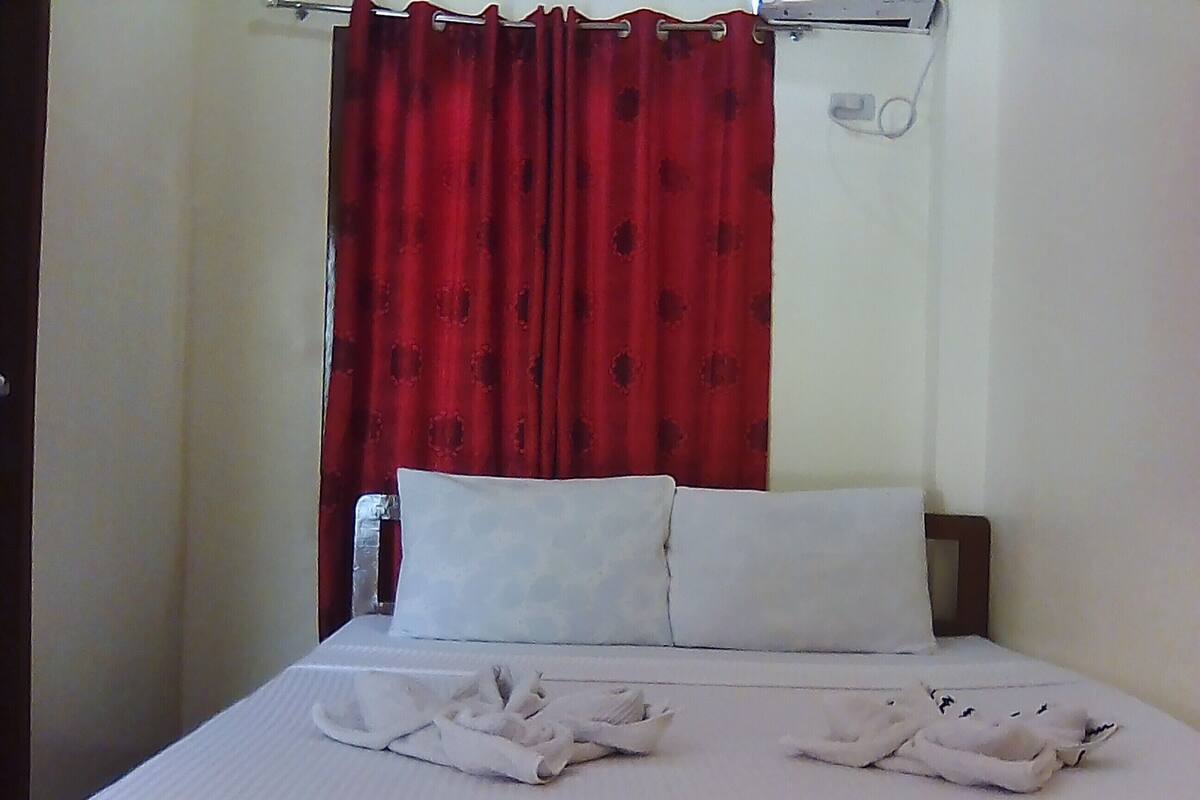 A bed with white linens and two pillows, a red curtain, and a small bedside table with a lamp.
