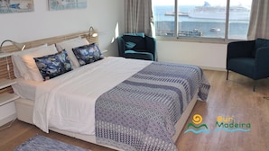Um quarto com uma cama, uma cadeira, uma janela com vista para um navio de cruzeiro e obras de arte na parede.