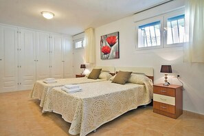 A bedroom with two beds, a nightstand with lamps, a painting, and a window with blinds. - Villa Calalga - PlusHolidays (Calpe)