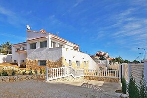 A two-story house with a white exterior and a stone base, a white fence, and a clear blue sky. - Villa Calalga - PlusHolidays (Calpe)