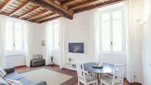A living room with a sofa, a dining table with chairs, a TV mounted on the wall, and large windows with curtains. - Rome, Trastevere, charming apartment for 4 (Rome)