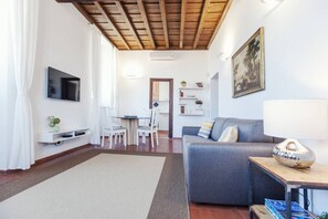 Ein Wohnzimmer mit einer Couch, einem an der Wand befestigten Fernseher, einem Essbereich mit Stühlen und einer Küche im Hintergrund.