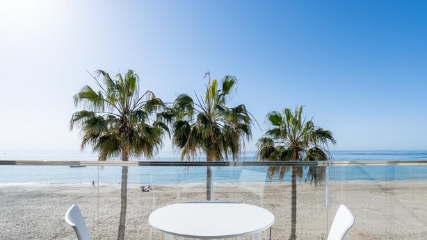 Un balcon avec une table blanche et deux chaises blanches, donnant sur une plage bordée de palmiers et sur l’océan.