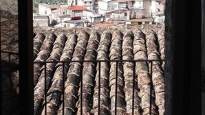 Vista a través de una ventana con celosía metálica, mostrando un pueblo con edificios y un paisaje montañoso al fondo.