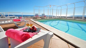 Un espace aménagé au bord de la piscine, avec des chaises longues, une table sur laquelle sont posées une serviette et des fruits, et une vue dégagée sur l’océan.