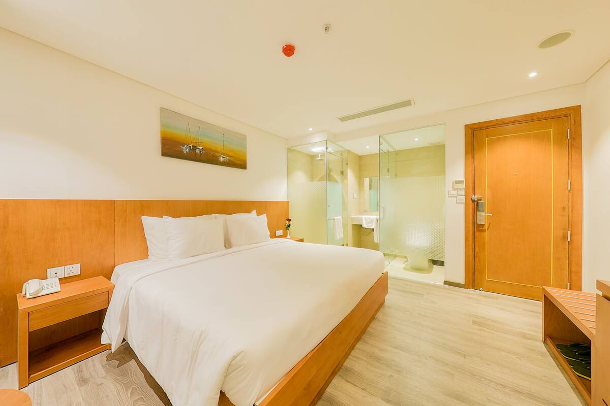 Habitación de hotel con una cama grande, cabecera de madera, mesita de noche con un teléfono, un cuadro en la pared y zona de ducha con mampara de vidrio.