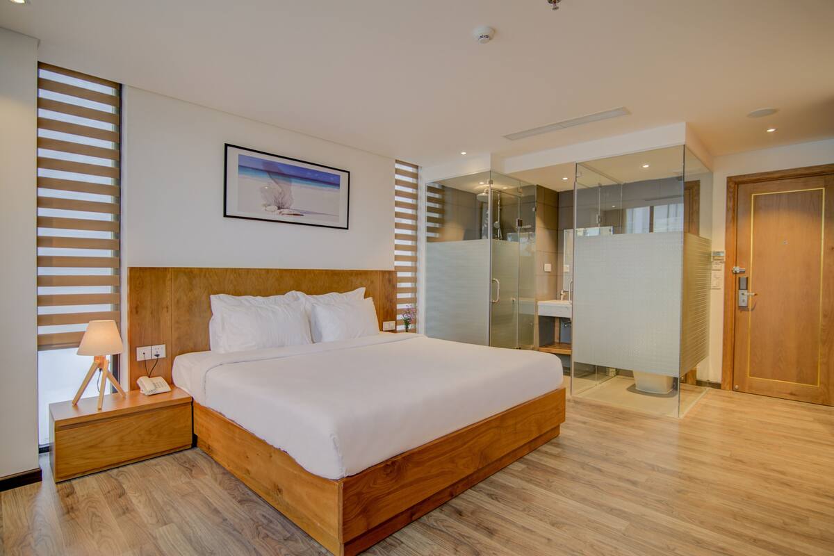 Una habitación de hotel moderna con una cama grande, una mesita de noche con una lámpara, un cuadro en la pared y un separador de vidrio que divide la habitación del baño.