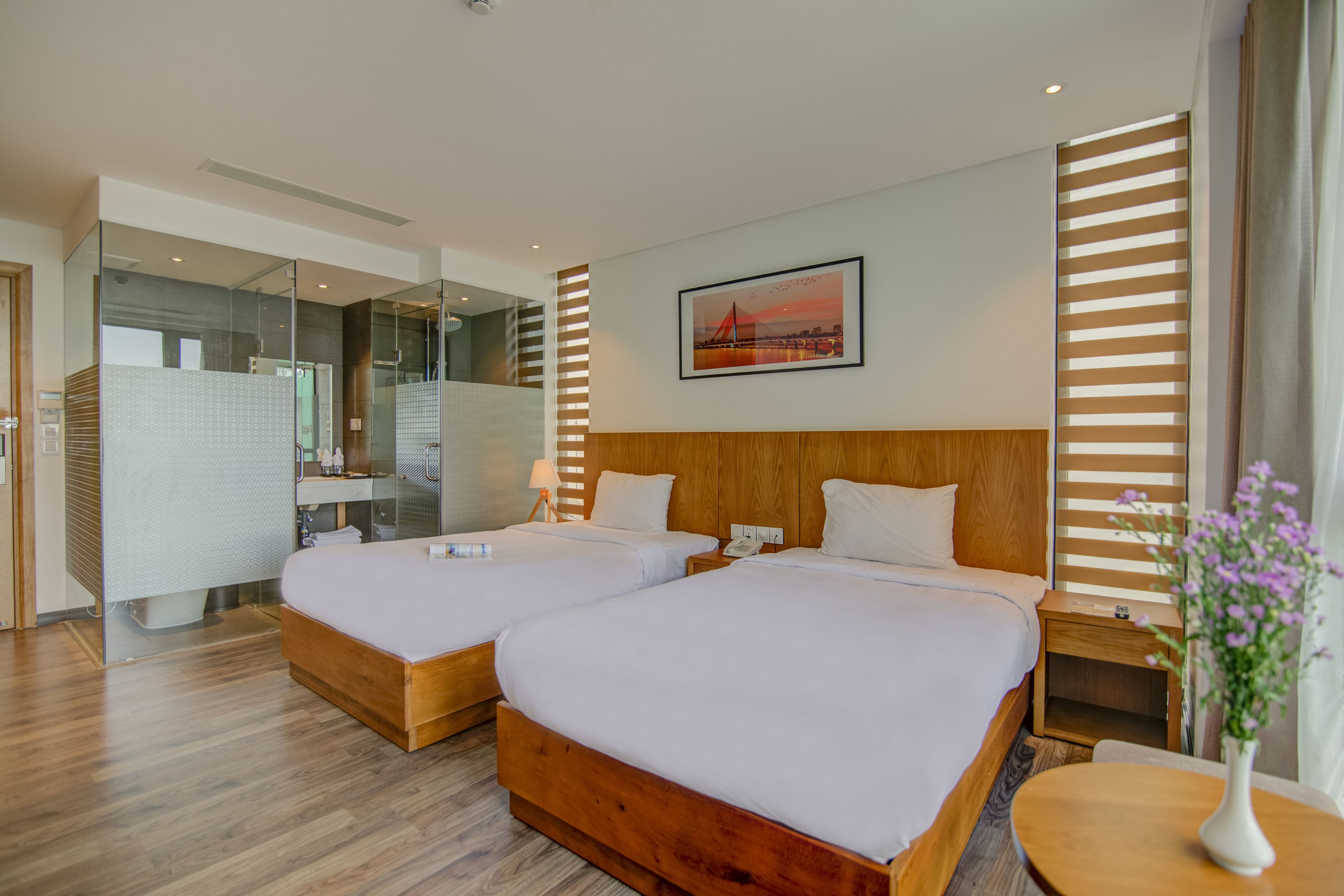Habitación de hotel con dos camas, cabecera de madera, mesita de noche con lámpara, una pequeña mesa con un jarrón de flores y un cuadro en la pared.