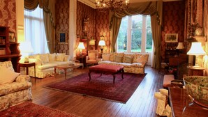 Ein geräumiges Wohnzimmer mit einem großen roten Teppich, verschiedenen Sitzmöglichkeiten, darunter eine Couch und Sessel, sowie einem Kronleuchter.