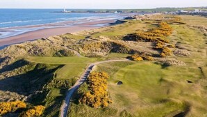 Eine Vogelperspektive eines Küsten-Golfplatzes mit einem Leuchtturm in der Ferne, einem gewundenen Weg und einem Strand.