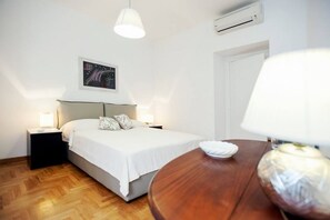 Une chambre à coucher comprenant un lit, des tables de chevet, une table ronde en bois, une lampe suspendue, un appareil de climatisation et un tableau accroché au mur.