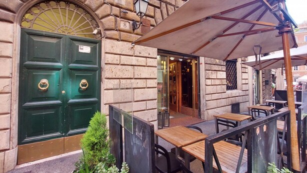 A building with a green double door, outdoor seating with tables and chairs, and a large umbrella providing shade. - Rome, near Piazza di Spagna, modern and spacious apartment for 8 (Roma)