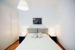 A bedroom with a bed, two bedside tables, a nightstand, a wall-mounted artwork, and a ceiling light fixture.
