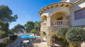 Un área de piscina con suelo de piedra, mobiliario exterior y un edificio con arcos y balcón.