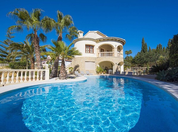 En stor, vit villa med en swimmingpool, omgiven av palmer och annan frodig vegetation.
