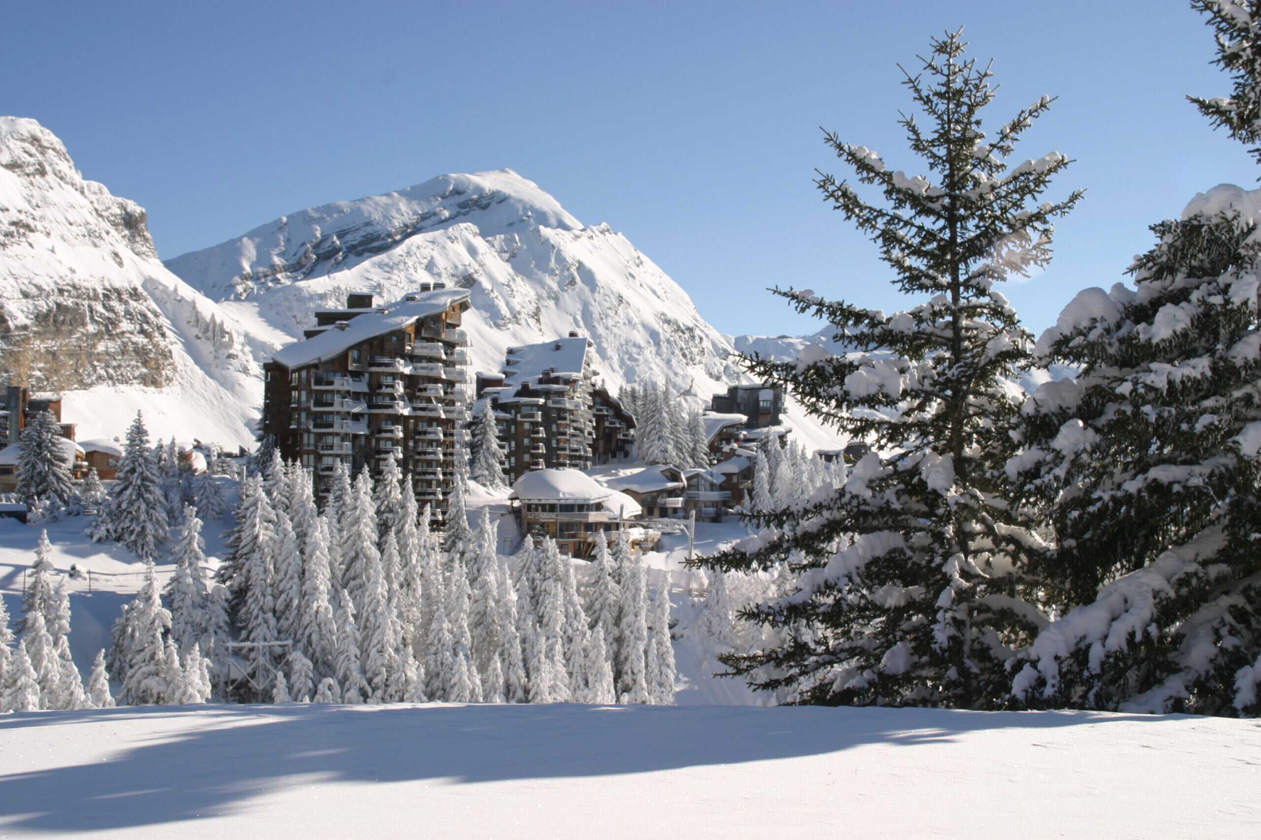 A snow-covered mountain resort with multiple buildings, including a large lodge with balconies, surrounded by snow-laden trees and a clear blue sky.