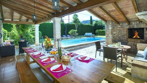 An outdoor dining area with a long wooden table set for a meal, overlooking a swimming pool and a stone fireplace. - VILLA VOLANTI - 10 MIN WALK TO THE OLD TOWN (Pollensa)