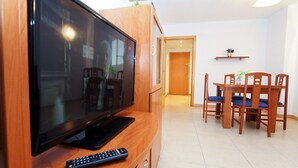 Ein Wohnzimmer mit Flachbildfernseher, Fernbedienung, Esstisch mit Stühlen und einer kleinen Topfpflanze auf einem Regal.