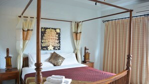 A four-poster bed with a wooden frame, white linens, a red quilt, and a decorative pillow. There are two bedside tables with lamps, and a large tapestry hangs on the wall. - Nirvana Guesthouse (Koh Tao)