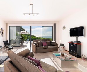 Een moderne woonkamer met een bank, salontafel en televisie. Er is een glazen deur naar een balkon met uitzicht op groen.