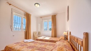 A hotel room with two beds, a window with curtains, a bedside table with a lamp, and a wall clock. - Villa Casita - PlusHolidays (Calpe)