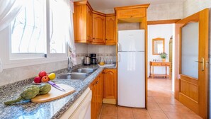A kitchen with wooden cabinets, a stainless steel sink, a white refrigerator, and a countertop with fruits and vegetables. - Villa Casita - PlusHolidays (Calpe)