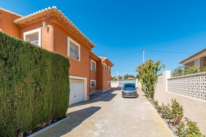 A residential street with a car parked in front of a house with a tiled roof and a well-maintained hedge. - Villa Casita - PlusHolidays (Calpe)