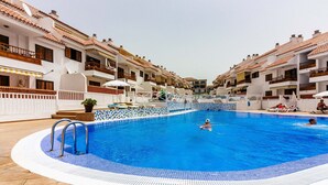 En swimmingpool omgiven av vita byggnader med balkonger, ett kaklat poolområde och en klar himmel.