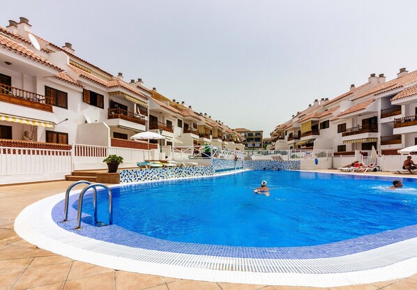 Ein Swimmingpool, umgeben von weißen Gebäuden mit Balkonen, einer gefliesten Terrasse und blauem Himmel.