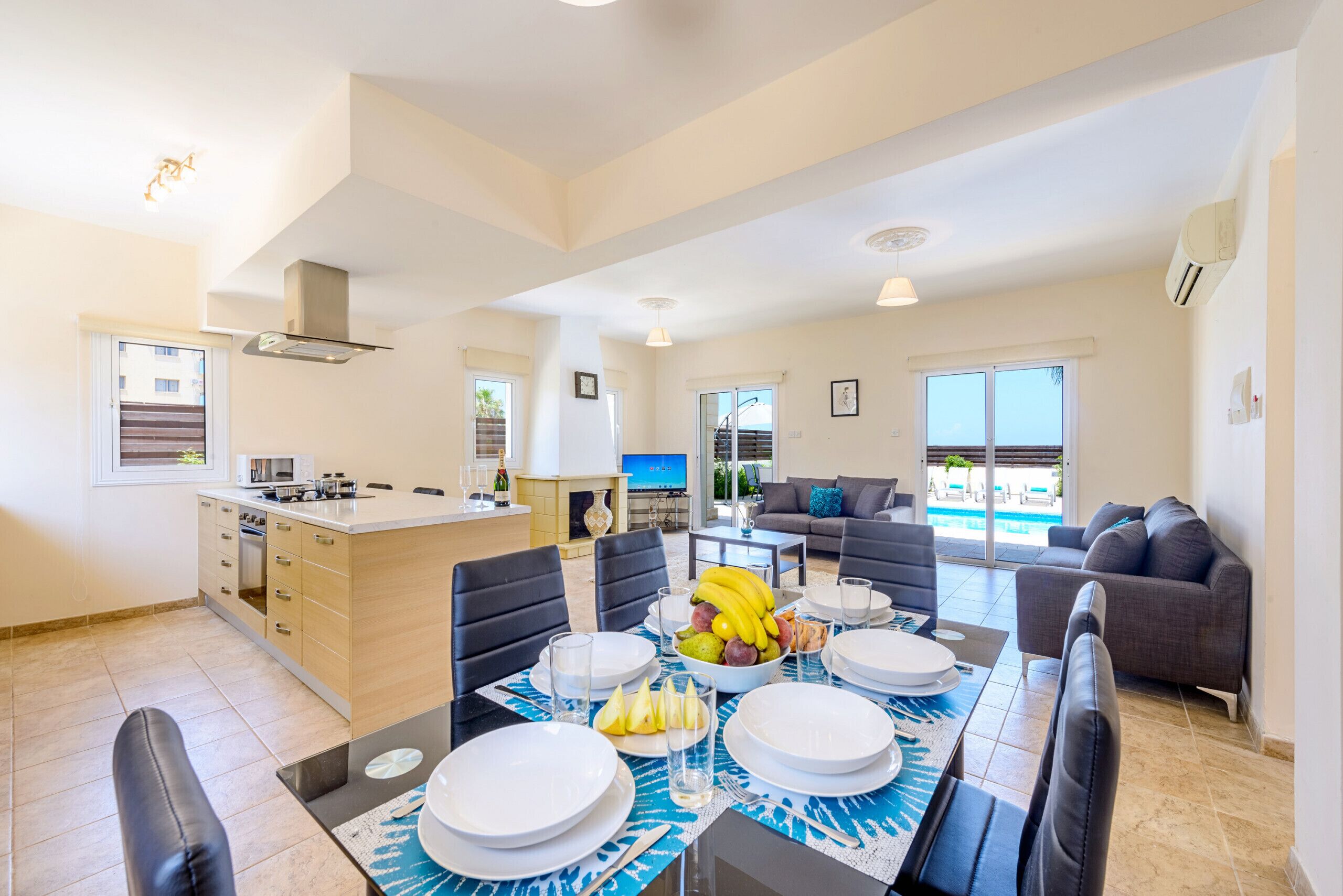 A modern kitchen and dining area with a large island, dining table set for six, and a living area with a sofa and a view of a pool.